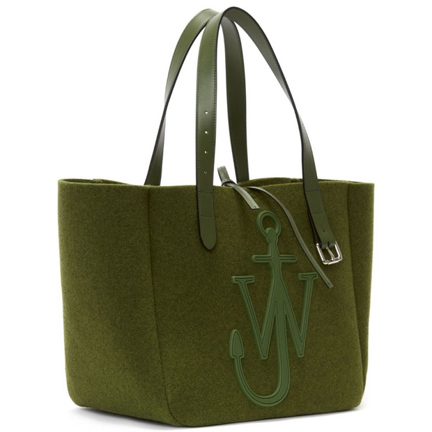 Angled side view of a polyester blend khaki tote with a leather tie closure. The tote has matching leather straps, one of which has a silver buckle hanging from it. A large leather brand logo is featured on the front. The tote is shown in its entirety against a white background.