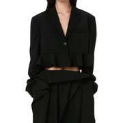 Image: A model wearing a black cropped blazer with a ruffle hem, shown from the chin to mid-thigh, against a white background.